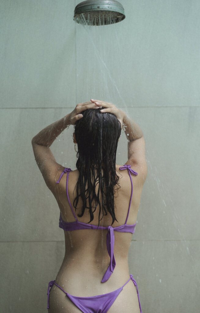 Back view of unrecognizable young sensual female in bikini touching dark wet hair while taking shower in bathroom